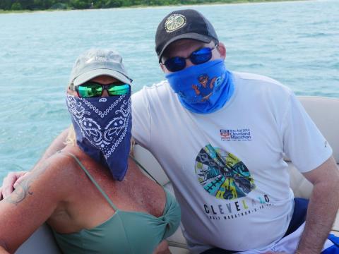 Two people on a boat, wearing sunglasses and bandanas over their faces, with water in the background.
