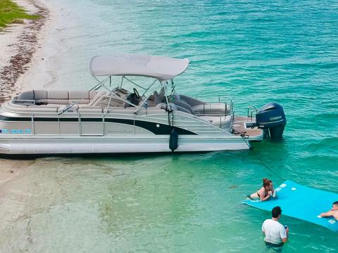 Pontoon boat docked on sandy shore with people swimming nearby.