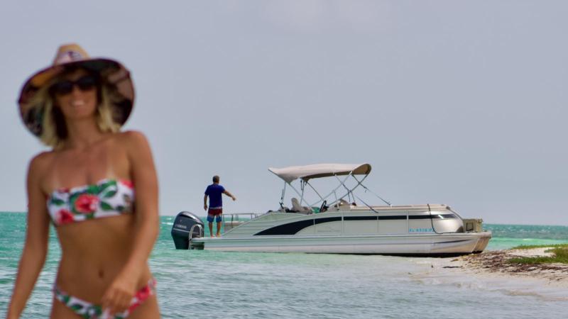 Woman in floral bikini and hat on beach, boat in background on water.