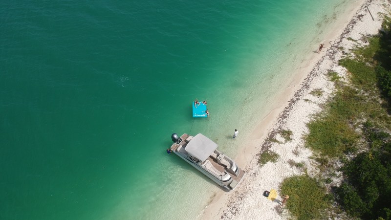 Aerial view of a motorboat and people lounging on a float near a sandy beach with turquoise water.