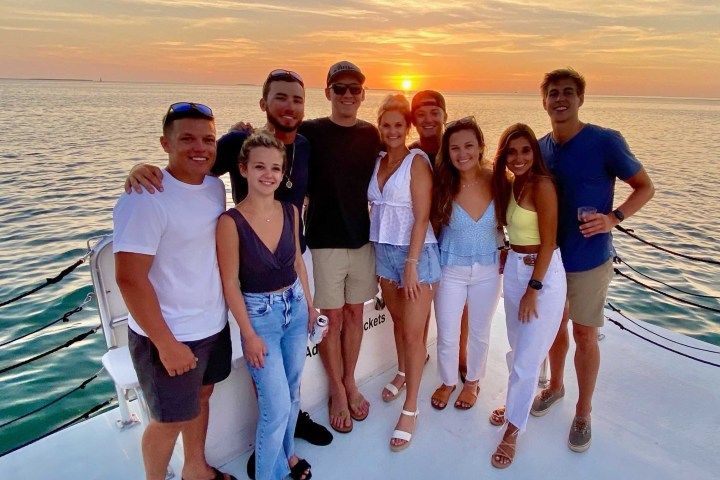 Group of nine people posing on a boat with a sunset in the background.